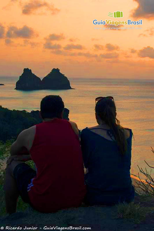 Imagem de casal sentados no mirante admirando o pôr do sol, na Praia do Boldro, em Fernando de Noronha, Pernambuco, Brasil.